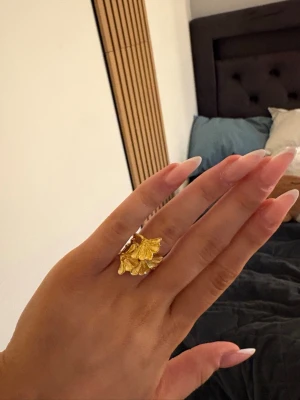 Guldfärgad ring med blomdetalj - Unik guldfärgad ring med en stor, detaljerad blomma framtill. Ringen har en öppen design med en tunn, grenliknande ringband som ger en naturlig och bohemisk känsla. Perfekt för dig som vill sticka ut med accessoarer som har en cool och kreativ touch.