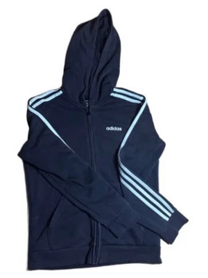 Mörkblå Adidas hoodie med dragkedja - Snygg mörkblå hoodie från Adidas med klassiska ljusblå ränder längs ärmarna och Adidas-logga på bröstet. Hoodien har dragkedja framtill, huva och ribbade muddar. Perfekt för en sportig och avslappnad stil. Materialet känns mjukt och bekvämt.