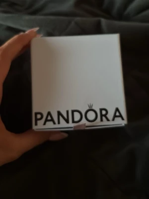 Pandora smyckesask i vitt - Säljer en snygg och stilren smyckesask från Pandora i vit kartong med svart inredning och rosa banddetalj. Perfekt för att förvara eller ge bort smycken. Locket har Pandoras logga i svart och insidan har silkespapper med logotypmönster.