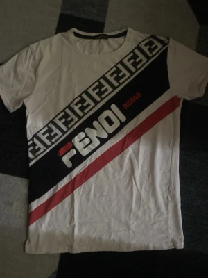Vit Fendi-inspirerad t-shirt med tryck - Säljer en vit t-shirt från Fendi med diagonal logga och texten 'ROMA' framtill. T-shirten har korta ärmar, rund hals och coola svarta, röda och vita detaljer samt ett grafiskt mönster över bröstet. Perfekt för dig som gillar streetwear och statement-plagg.