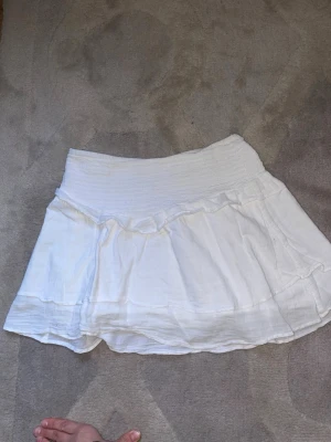 Vit kortkjol från Gina Tricot - Söt vit kortkjol från Gina Tricot med bred smock i midjan och volangdetaljer. Kjolen är gjord i ett lätt och luftigt bomullstyg, perfekt för varma dagar. Enkel att matcha med olika toppar för en fräsch look. Kan gå ner i pris