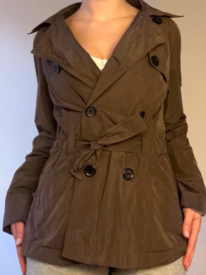 Brun trenchcoat med knytskärp🤎 - Stilren brun trenchcoat med dubbelknäppning och stora svarta knappar. Jackan har ett matchande knytskärp i midjan och klassisk krage. Tillverkad i ett lätt tyg som ger en avslappnad look. Perfekt för dig som vill ha en tidlös och trendig jacka.🤎