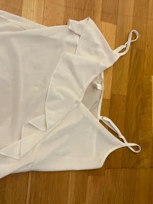 Vit linnetopp med volangdetalj - Snygg vit linnetopp med tunna axelband och volangdetalj framtill. Perfekt för varma dagar och enkel att styla med jeans eller kjol. Tunn och luftig känsla, passar dig som gillar minimalistisk stil.