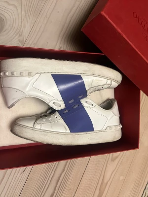 Valentino open sneakers - Säljer ett par vita sneakers från Valentino med en bred blå med lite lila nyans remsa över mitten. Skorna har klassisk låg profil, vita snören och nitar längs sulan. Tillverkade i läder och levereras i originalkartong.