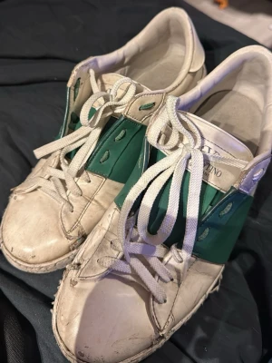 Vita och gröna sneakers från Valentino - Säljer ett par snygga sneakers från Valentino i vitt läder med grönt band över mitten och vita skosnören. Klassisk låg modell med rund tå och diskret logga på plösen. Perfekta för dig som gillar exklusiva märken och stilren design lite slitiga men pris går att diskutera 