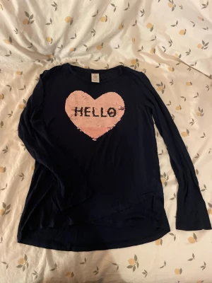 Svart långärmad topp med paljetthjärta barn st 134/140 - Snygg svart långärmad topp med ett stort rosa hjärta i paljetter på bröstet och texten 'HELLO' i svart. Rund halsringning och mjukt material som känns skönt mot huden. Perfekt för dig som gillar statement-plagg och glitter.