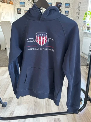Mörkblå hoodie från Gant - Snygg mörkblå hoodie från Gant med broderad logga och texten 'American Sportswear' på bröstet. Klassisk känguruficka framtill och huva. Tillverkad i mjukt bomullsmaterial, perfekt för en avslappnad och sportig stil.