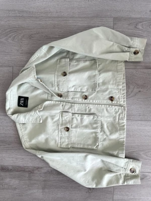 Ljusgrön jeansjacka från ZARA - Trendig ljusgrön jeansjacka från ZARA med två stora bröstfickor och snygga bruna knappar framtill. Klassisk krage och raka ärmar med knapp vid ärmslut. Perfekt för dig som vill ha en stilren och modern look.