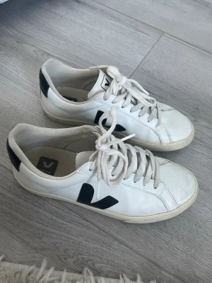 Vita Veja sneakers med svart detalj - Snygga vita sneakers från Veja med klassisk svart V-logga på sidan och svarta detaljer vid hälen. Skorna har vita snören och en beige sula. Tillverkade i läder med en stilren och tidlös design som passar till det mesta.