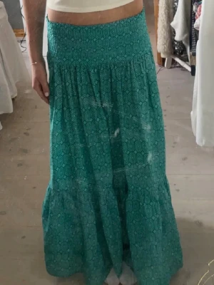 Grön långkjol med grafiskt mönster - Snygg grön långkjol med grafiskt mönster och smock i midjan. Kjolen har en luftig och flowy passform, perfekt för varmare dagar. Tillverkad i ett mjukt bomullstyg som känns skönt mot huden.