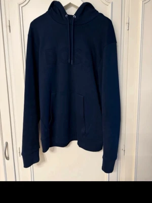 Mörkblå hoodie från BOSS - Snygg mörkblå hoodie från BOSS med broderad logga på bröstet. Klassisk modell med huva och dragsko samt stor känguruficka framtill. Tillverkad i mjukt bomullsmaterial som känns skönt mot huden. Perfekt för en avslappnad och trendig look.