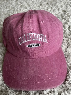 Rosa keps med California-brodyr - Säljer en rosa keps med böjd skärm och broderad text 'CALIFORNIA' och 'WEST COAST' framtill i vitt. Kepsen har klassisk passform och är tillverkad i bomullstyg. Perfekt accessoar för en avslappnad och trendig look.