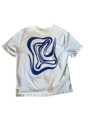 Vit t-shirt från J.lindeberg - Vit t-shirt från j.lindeberg i bomull med ett stort blått grafiskt tryck på ryggen och en liten blå textdetalj på bröstet. Klassisk passform och rund halsringning. Perfekt för dig som gillar minimalistisk stil med en twist. En liten fläck på magen/genomskinlig syns knappt