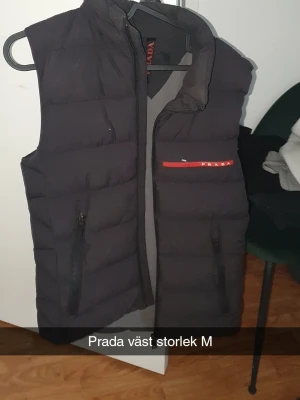 Svart dunväst från Prada - Snygg svart dunväst från Prada med diskret röd logga på bröstet och dragkedjor framtill. Västen har en klassisk, quiltad design och står ut med sin minimalistiska look. Perfekt för lager-på-lager och streetstyle. Litet hål under loggan som inte är synligt på långt håll dragkedjan går inte att stänga men enkelt att laga hos kunnig, västen är äkta!