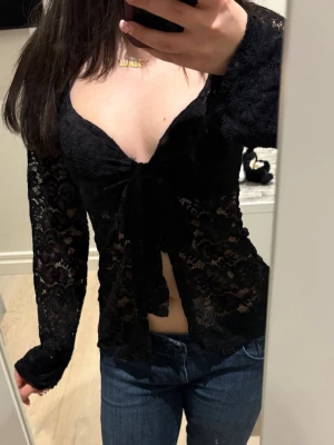 Svart spetsblus med knyt framtill - Säljer en svart långärmad topp i genombruten spets med djup urringning och knyt framtill. Modellen är figurnära och har transparenta ärmar och kropp. Perfekt för dig som gillar trendiga och feminina plagg.