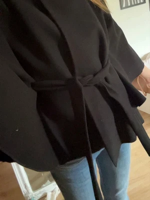 Svart cape-kappa med knytband - Stilren svart cape-kappa med knytband i midjan och hög krage. Kappan har en dekorativ knapp upptill och vida ärmar för en trendig siluett. Perfekt för dig som vill ha en elegant och modern look. Materialet ser ut att vara mjukt och tjockt, troligen ullblandning.