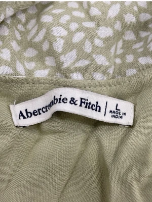 Grön omlottklänning Abercrombie & Fitch i naturmaterial  - Ljusgrön omlottklänning från Abercrombie & Fitch med vitt abstrakt mönster. Klänningen har knyt i midjan och volangdetaljer nedtill. Tillverkad i 100% cellulosa , perfekt för sommarens varma dagar.