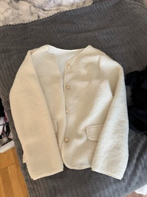 Krämvit teddyjacka med knappar - Mysig krämvit teddyjacka med rund halsringning och fyra knappar framtill. Jackan har två fickor och är gjord i fluffigt teddy-material som ger en soft vibe. Perfekt för lager på lager och chill stil.