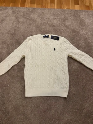 Vit kabelstickad tröja Polo Ralph Lauren - Kabelstickad vit tröja från Polo Ralph Lauren med klassisk rund halsringning och den ikoniska broderade loggan på bröstet. Tröjan har ribbade muddar vid ärmslut och nederkant. Perfekt för en clean och stilren look.