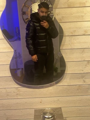 Svart dunjacka från Moncler - Snygg svart dunjacka från Moncler med glansig finish och huva. Jackan har lång ärm, dragkedja framtill och två sidofickor. Perfekt för kalla dagar och ger en riktigt modern look.  JACKAN ÄR INTE I PERFEKT SKICK. Finns ett litet litet hål i jackan men det är inget man märker för en extra summa kan jag fixa jackan innan du köper den. Det är bara att skriva vid funderingar är tillgänglig 24/7 och svarar alltid så snabbt som möjligt.