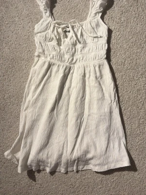 Vit sommarklänning med knyt - Söt vit klänning i lätt, skrynkligt tyg med breda axelband och knyt framtill. Perfekt för varma dagar och har en avslappnad passform. Midjan är markerad med sömmar för en feminin siluett. Aldrig använd men har ej kvar prislapp.
