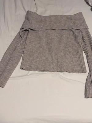 Grå off shoulder stickad tröja H&M - Säljer en ribbstickad grå off shoulder-tröja från H&M. Tröjan har långa ärmar och en bred, nedvikt krage som ger en snygg och avslappnad look. Perfekt att matcha med jeans eller kjol för en trendig outfit.