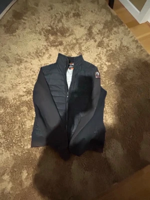 Cardigan PJS - Stilren svart cardigan från pjs med hög krage och dragkedja framtill. Västen har ett emblem på axeln och quiltad design på framsidan. Insidan är ljusgrå och materialet är lätt och smidigt, perfekt för lager-på-lager. Klassisk look som funkar till det mesta.