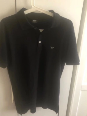 Svart piké från Armani - Klassisk svart pikétröja från Armani med korta ärmar och liten logga på bröstet. Tröjan har krage och knäppning med två knappar framtill. Perfekt för en clean och stilren look.