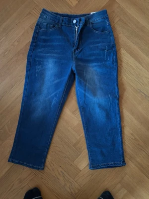 Blå raka trekvartslånga jeans med slitningar - Säljer ett par blå trekvartslånga jeans med raka ben och diskreta slitningar framtill. Jeansen har klassisk femficksdesign, bälteshällor och dragkedjegylf med knapp. Materialet är denim i bomull och passformen är relaxed.