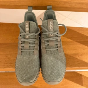 Grå Adidas sneakers med mesh - Snygga grå sneakers från Adidas med stickad mesh-ovandel och matchande grå sula. Skorna har klassiska snörningar och Adidas-logga på plösen. Perfekta för dig som gillar stilrena och sportiga sneakers med modern design.