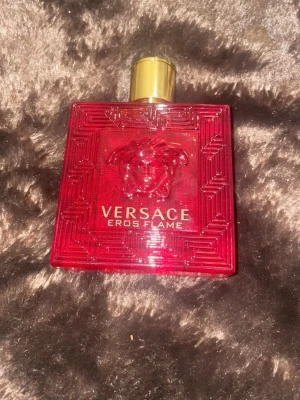 Versace Eros Flame röd parfym - Versace Eros Flame parfym i en snygg, fyrkantig röd glasflaska med guldfärgat lock och Medusa-detalj. Flaskan har ett elegant mönster och tydlig logga framtill. Perfekt för dig som gillar exklusiva och stilrena dofter.