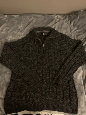 Mörkgrå stickad tröja med halv dragkedja - Mysig mörkgrå stickad tröja med halv dragkedja framtill och hög krage. Tröjan har ett diskret melerat mönster, långa ärmar och ribbstickade muddar vid ärmslut och nederkant. Perfekt för kalla dagar och enkel att matcha med jeans.