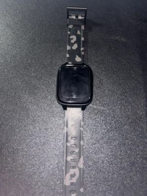 Svart smartklocka med camo-armband - Snygg smartklocka med fyrkantig svart urtavla och ett grått kamouflagemönstrat silikonarmband. Modern design som passar till det mesta och känns bekväm på handleden. Perfekt för dig som vill ha koll på notiser och hälsa direkt på handleden. Den är helt ny bara använt 3 gånger och det finns en sim kort plats och det kommer en snabb ladare