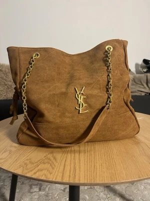 Brun mocka handväska från YSL - Snygg handväska i brun mocka från YSL med guldfärgade kedjehandtag och ikoniska YSL-loggan framtill. Väskan har en mjuk, slouchy form och detaljer i guldmetall. Perfekt för dig som gillar lyxiga accessoarer med vintagekänsla.
