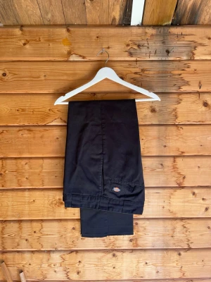Svarta chinos från Dickies - Säljer ett par svarta chinos från Dickies med klassisk rak passform och diskret logga på bakfickan. Byxorna har en stilren look och är tillverkade i ett slitstarkt bomullsmaterial. Perfekta för dig som gillar enkel och clean stil.