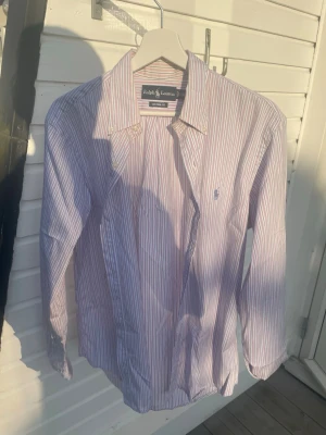 Randig skjorta från Ralph Lauren - Klassisk skjorta från Ralph Lauren i rosa med tunna blå och vita ränder. Skjortan har lång ärm, krage och knappar framtill. Tillverkad i 100% bomull för en skön och luftig känsla. Perfekt för en stilren och avslappnad look.