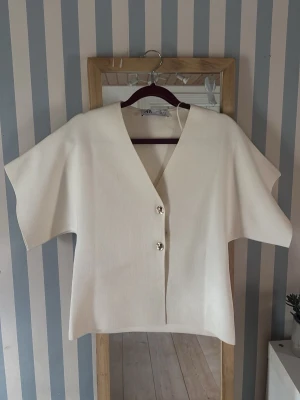Vit v-ringad blus med knappar - Stilren vit blus med v-ringning och två dekorativa silverknappar framtill. Modellen har vida, korta ärmar och rak passform. Materialet känns mjukt och har en lätt struktur. Perfekt för dig som gillar minimalistisk och clean stil.