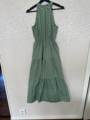 Grön prickig långklänning - Snygg grön långklänning med vita prickar och markerad midja. Klänningen har ärmlös design, rundad halsringning och faller i flera lager för en luftig känsla. Perfekt för varma dagar och somriga tillfällen.