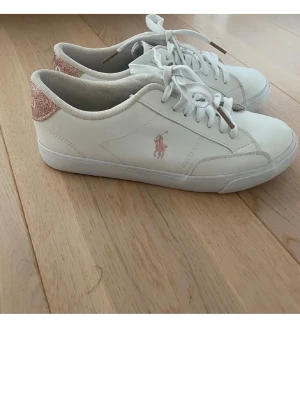 Vita Polo Ralph Lauren sneakers SÅLDA! - Snygga vita sneakers från Polo Ralph Lauren med rosa glitterdetalj på hälen och klassisk logga i rosa på sidan. Skorna har lågt skaft, vita snören och är tillverkade i slätt syntetmaterial. Perfekta för en trendig och sportig look. OBS! DOM ÄR SÅLDA!
