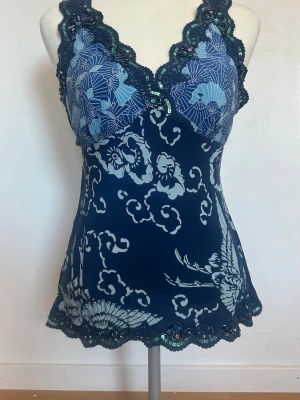 Mörkblå blommig linnetopp med spets och paljetter - Mörkblå linnetopp med ljusblå blommigt mönster, v-ringad hals och dekorativ spetskant med paljetter. Toppen har knyt i ryggen och är ärmlös, vilket ger en feminin och trendig look. 