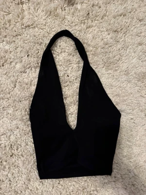 Svart halterneck topp med djup urringning - Snygg svart halterneck topp med djup v-ringning och croppad passform. Toppen är ärmlös och har en enkel, stilren design som passar perfekt till många olika looks. Tillverkad i mjukt material som sitter skönt mot kroppen.