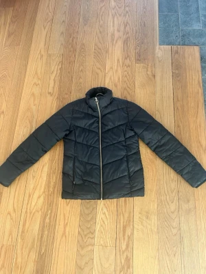 Svart pufferjacka med dragkedja - Snygg svart pufferjacka med hög krage och dragkedja framtill. Jackan har ett quiltat mönster i chevron-stil och två sidofickor med dragkedja. Perfekt för kalla vinterdagar och ger en clean look till din outfit. Den har även en avtagbar luva och pälskant så kan nästan se ut som två olika jackor🥰