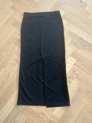 Svart långkjol med slits - En stilren svart långkjol i mjukt material med rak passform. Kjolen har en diskret slits baktill som ger extra rörelsefrihet. Perfekt att matcha med en crop top eller stickad tröja för en trendig look.