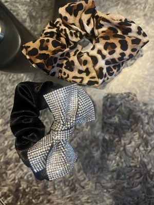 Två scrunchies med rosett - Säljer två scrunchies: en i glansigt svart tyg med stor rosett täckt av silvriga strass, och en i satinliknande material med leopardmönster och rosett. Perfekta för att piffa till håret med lite extra attityd.