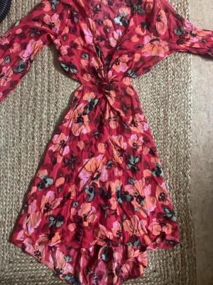 Blommig omlottklänning från H&M - Snygg blommig omlottklänning från H&M i rött, orange och grönt. Klänningen har lång ärm, v-ringning och volangdetaljer nertill. Perfekt för sommarens alla tillfällen och har en feminin passform med knyt i midjan.