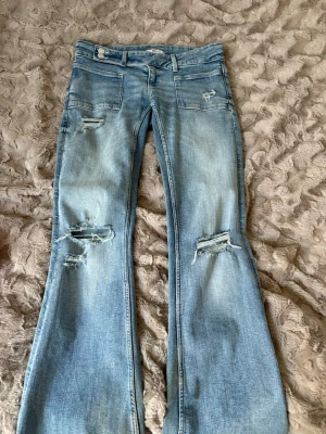 Ljusblå jeans  - Säljer ett par ljusblå bootcut jeans från Gina Tricot med slitningar och hål på benen för en trendig look. Jeansen har två framfickor och är tillverkade i klassisk denim. Perfekta för dig som gillar en avslappnad och cool stil.