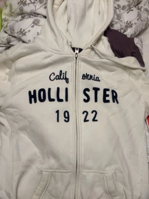 Vit hoodie från Hollister med tryck - Säljer min brorsas ex vita hoodie från Hollister med dragkedja framtill och stor huva. Broderat mörkblått tryck med texten 'HOLLISTER 1922 California' på bröstet. Två fickor framtill och ribbade muddar. Mjuk och skön bomullsblandning, perfekt för en avslappnad stil. Någon fläck men det går nog att få bort i tvätten om man försöker. Men det påverkar iaf inte helhetsintrycket.