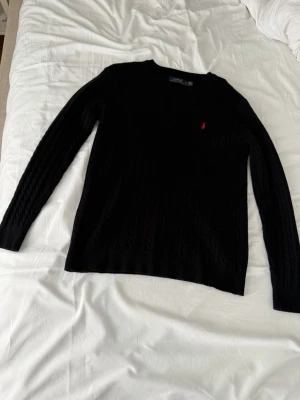 Svart kabelstickad tröja Polo Ralph Lauren - Kabelstickad svart tröja från Polo Ralph Lauren med rund halsringning och den klassiska röda loggan broderad på bröstet. Tröjan har långa ärmar och ribbade muddar. Perfekt för dig som gillar stilrena och tidlösa plagg.