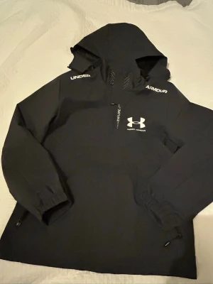Svart vindjacka från Under Armour - Snygg svart vindjacka från Under Armour med huva och dragkedja framtill. Jackan har vita logotyper på bröstet och axlarna samt elastiska muddar vid ärmsluten. Perfekt för blåsiga dagar och sportiga outfits.