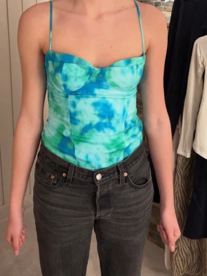 Blågrön body med tunna band - Hej!☀️Jag säljer en snygg body i blått och grönt med tie-dye mönster och tunna axelband. Toppen har markerade kupor och figurnära passform, perfekt för dig som vill sticka ut. Materialet känns mjukt och stretchigt, troligen polyester eller bomullsmix. Lite slitage (syns på sista bilden) men är inget man tänker på💕 Väldigt fin!💕💕